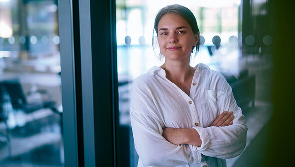 Eine junge Mitarbeiterin des Arbeitgebers ORAFOL lächelt freundlich in die Kamera. Sie steht vor einer Glastür in einem Verwaltungsgebäude und trägt eine lässige weiße Bluse, die Arme sind vor der Brust verschränkt. 
